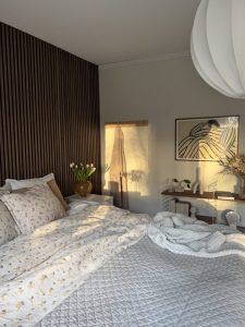 Sfeervolle slaapkamer met WoodUpp houten lattenwand achter het bed, een WoodUpp houten kapstok aan de muur en een WoodUpp smalle houten plank/console rechts. Zacht beddengoed, nachtkastje met vaas tulpen en warme avondzon die schaduwen op de wanden werpt met decoratieve accessoires.