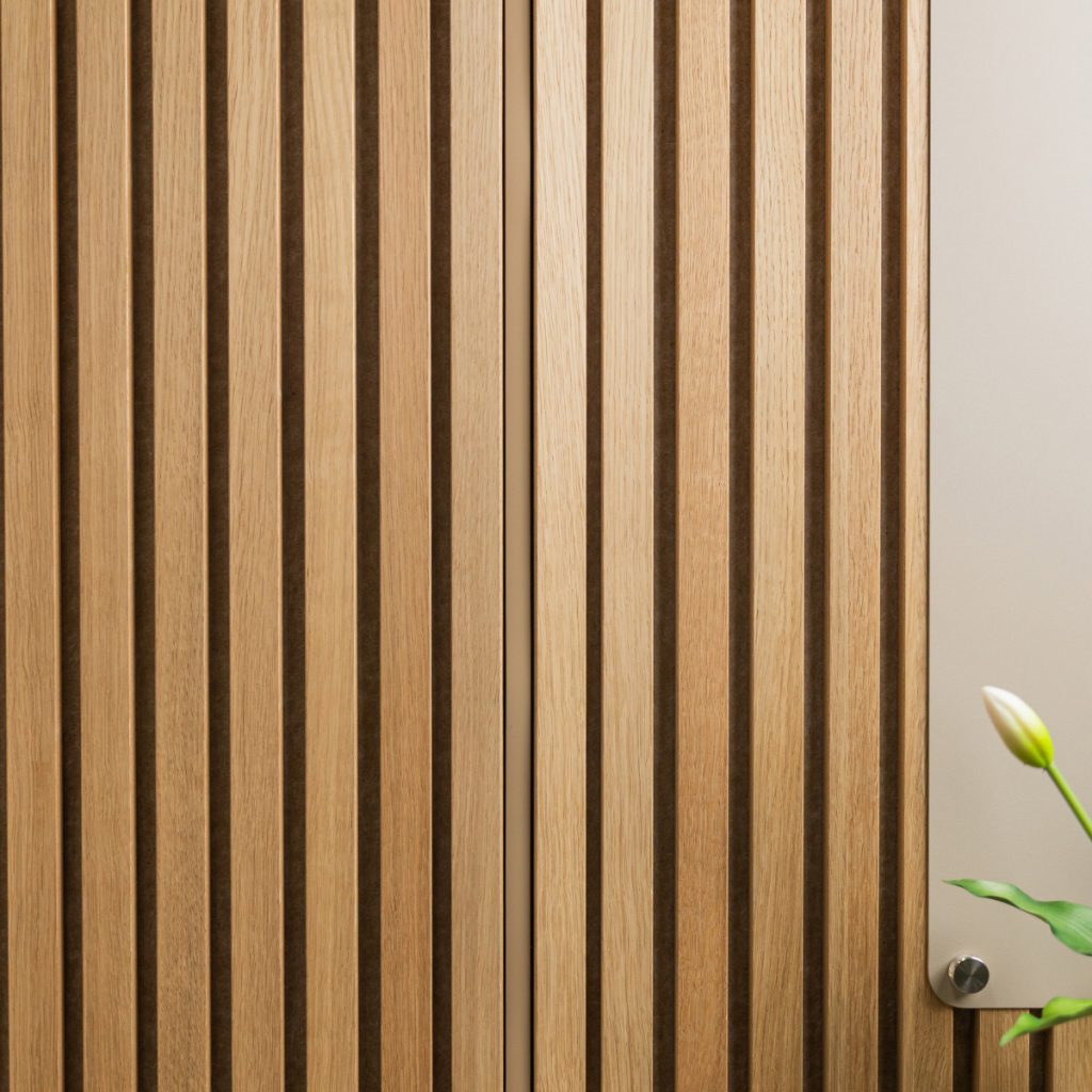 Close-up van een WoodUpp eiken lamellenwand: gelijkmatige, verticale houten latten met diepe donkere tussenruimtes. Rechts een beige WoodUpp montagepaneel met ronde metalen afstandhouder en een witte tulp zichtbaar. Strakke, natuurlijke afwerking die akoestische en decoratieve eigenschappen benadrukt en warme houttinten.