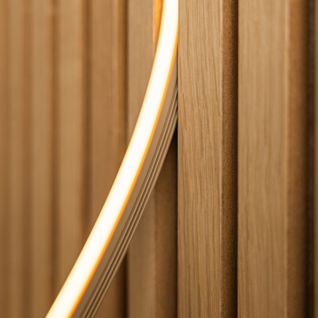 Close-up van een WoodUpp eiken lamellenwand met smalle, verticale houten lamellen en zichtbare nerf en fijne profilering. Een gebogen, warmwitte WoodUpp LED-neonstrip loopt schuin langs de lamellen en accentueert het reliëf, de warmte en de strakke, moderne afwerking van het interieur.
