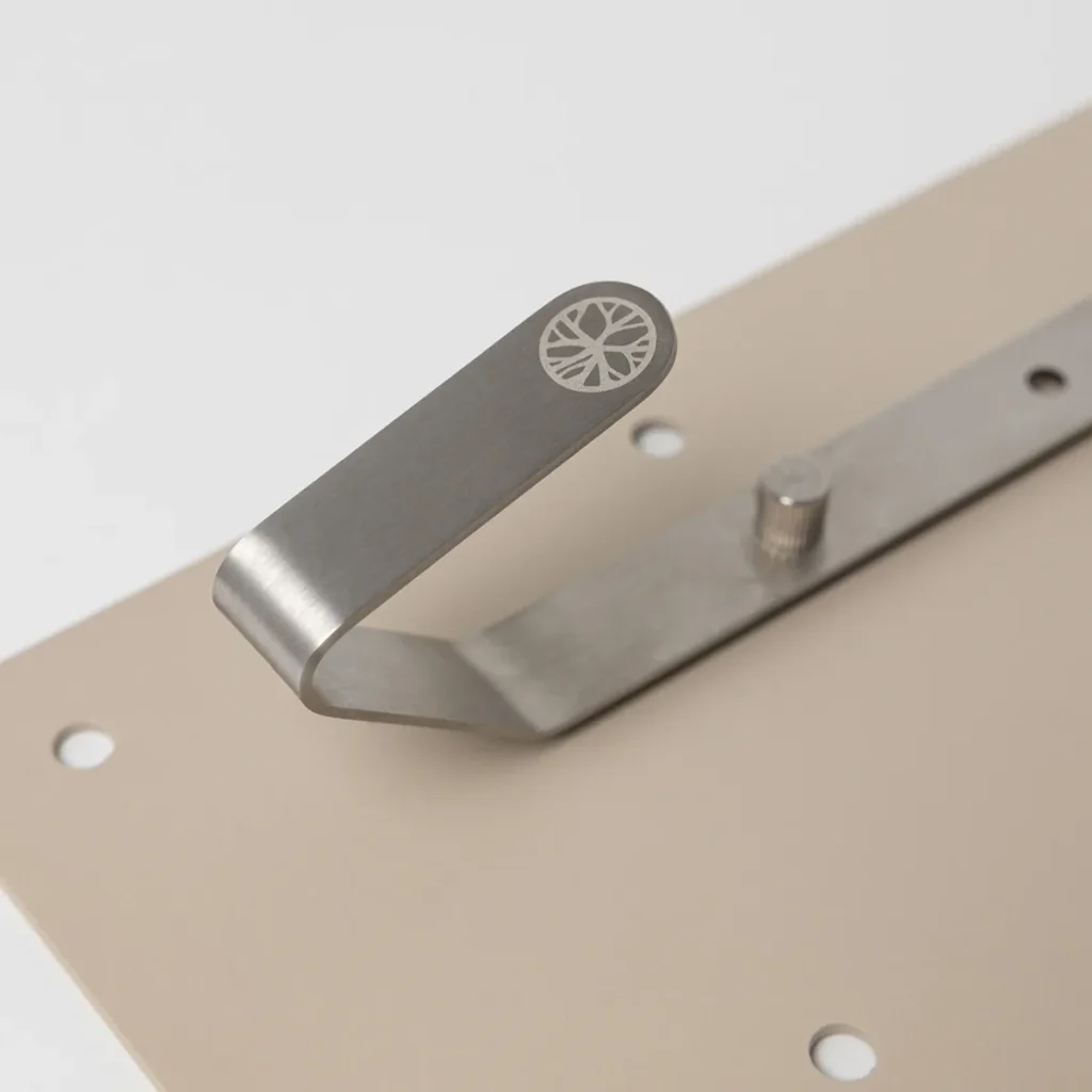 Close-up van een WoodUpp roestvrijstalen haak gemonteerd op een beige wandplaat; de slanke, gebogen metalen kapstokhaak heeft een geëtst WoodUpp-boomsymbool op het uiteinde en een verstelbare bevestigingspen, zichtbaar met montagegaten in de plaat. De matte, geborstelde finish accentueert het minimalistische, moderne ontwerp.