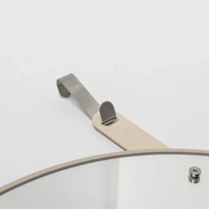 Close-up van een WoodUpp wandspiegel: ronde spiegel met beige metalen rand en brede leren ophangband, bevestigd aan een geborstelde RVS ophanghaak en schroef. Minimalistische bevestiging toont het stevige, elegante ontwerp van WoodUpp met hoogwaardige materialen en subtiele afwerking.