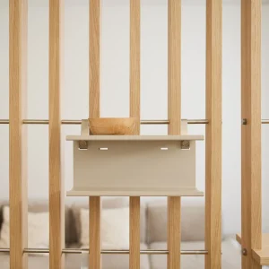 WoodUpp eiken lattenwand met daaraan een zandkleurige, wand‑gemonteerde opbergplank van WoodUpp, bevestigd met metalen stangen en subtiele clips; op de plank staat een kleine, handgesneden houten kom. Minimalistisch en functioneel roomdivider-ontwerp met naturel houtnerf, strakke lijnen en matte afwerking.