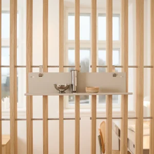 WoodUpp opbergpaneel met slanke, platte beige plank in neutrale tint, bevestigd aan verticale eikenlatten met metalen klemmen; erop een glanzende RVS-kom, een hoog cilindervormig RVS-vaasje en een klein gedraaid houten schaaltje — toont het modulaire, minimalistische opberg- en display-systeem van WoodUpp in een lichtrijke, Scandinavisch geïnspireerde ruimte.