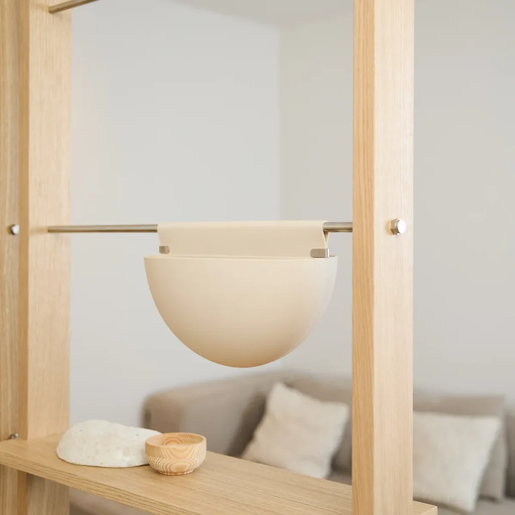 WoodUpp houten roomdivider/rek met verticale eiken stijlen en een metalen stang waaraan een beige, halfronde hangpot/organizer hangt. Op het houten plankje liggen een lichtgekleurde steen en een klein, rond WoodUpp houten kommetje, met een strakke, Scandinavische uitstraling en glanzende metalen bevestigingen.