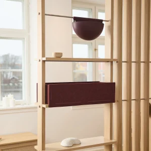 WoodUpp eikenhouten roomdivider met slanke verticale latten en smalle planken, gefotografeerd met een bordeauxrode vilten opbergbox die in het frame schuift en een gelijkgekleurde halfbolvormige hangpot. Kleine houten kom en witte decoratieve steen op plank maken het geheel sfeervol en functioneel.