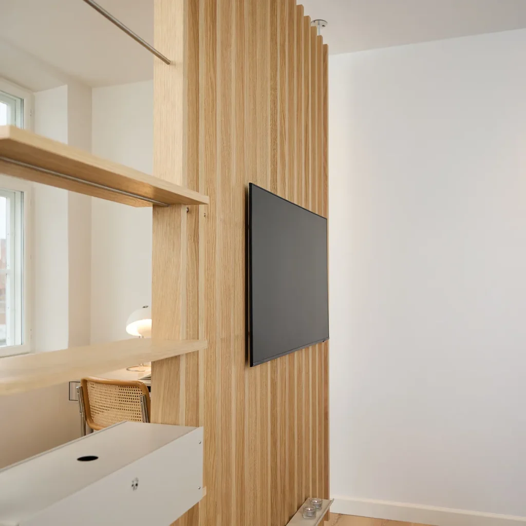 WoodUpp lamellenwand van licht eikenfineer met smalle verticale lamellen als roomdivider, inclusief geïntegreerde tv‑bevestiging met platte zwarte televisie. Aan de zijkant twee WoodUpp zwevende eiken planken zichtbaar, strak gemonteerd, ruimtebesparend en met natuurlijke houttextuur tegen een witte wand.