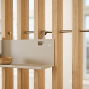 Close-up van WoodUpp houten lattenwand met verticale eikenhouten lamellen en een roestvrijstalen horizontale stang waaraan een zandkleurige metalen WoodUpp wandplank is opgehangen; op de plank staat een klein, geribd houten kommetje, zichtbaar het metalen ophanghaakje van het modulaire opbergsysteem en functioneel.