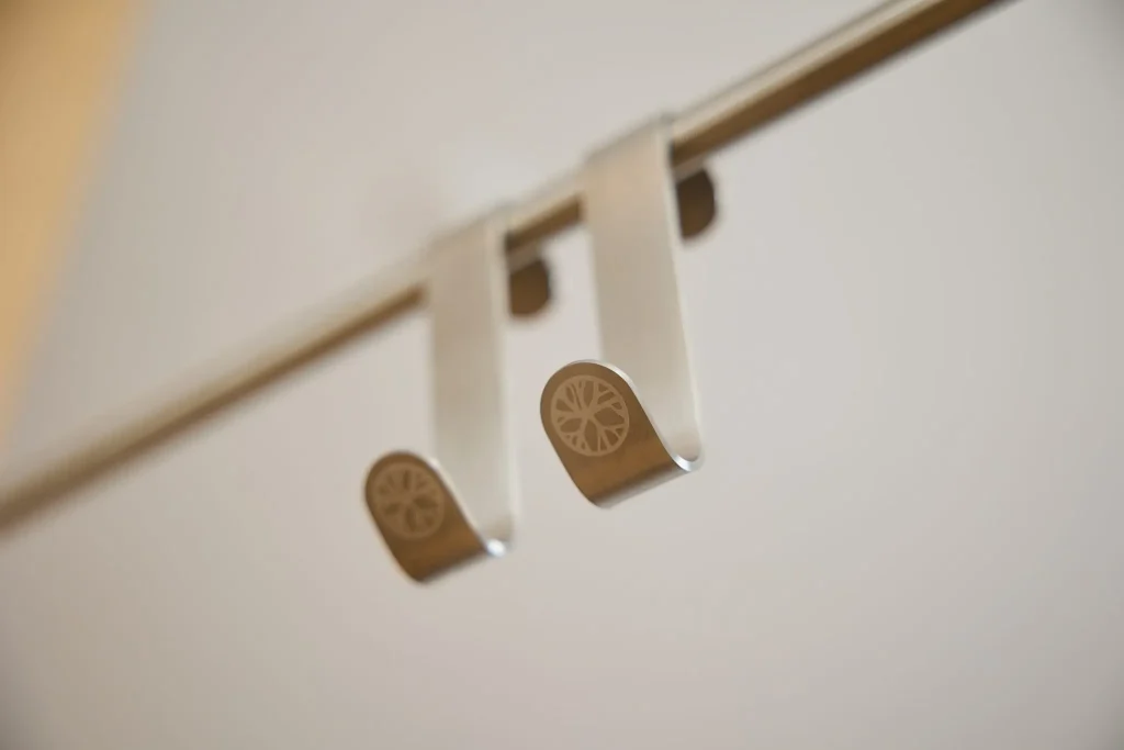 Close-up van twee WoodUpp over-deurhaken van geborsteld roestvrij staal, hangend aan een smalle metalen rail. Slanke, zilverkleurige, gebogen haken met gegraveerd WoodUpp-logo op de uiteinden; strak, functioneel ontwerp, ideaal voor het ophangen van kleding, handdoeken en accessoires in huis voor dagelijks gebruik.
