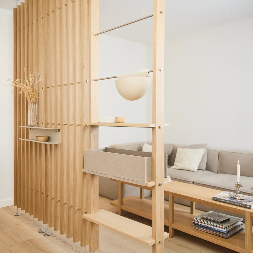 WoodUpp roomdivider van lichte eikenlatten met metalen stangen, ingebouwde houten planken en een schuifbare vilten opbergbox. Op de planken staan een vaas met droogbloemen en een klein schaaltje; het praktische, modulair opgebouwde WoodUpp-systeem deelt de woonkamer en biedt extra opbergruimte en sfeer.