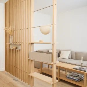 WoodUpp roomdivider van lichte eikenlatten met metalen stangen, ingebouwde houten planken en een schuifbare vilten opbergbox. Op de planken staan een vaas met droogbloemen en een klein schaaltje; het praktische, modulair opgebouwde WoodUpp-systeem deelt de woonkamer en biedt extra opbergruimte en sfeer.