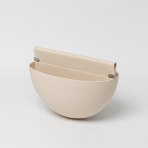 Een beige, halfrond opbergbakje van WoodUpp, ontworpen als accessoire. Het bakje heeft een gladde, matte afwerking die doet denken aan siliconen, en is voorzien van kleine metalen clips aan de zijkanten van de bevestigingsflap aan de achterzijde. Het staat tegen een neutrale lichte achtergrond.