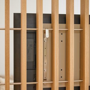 Close-up van een WoodUpp eiken lamellenwand met rechte, slanke latten en horizontale roestvrijstalen stangen; achter de lamellen is de achterkant van een tv zichtbaar, bevestigd aan een WoodUpp stalen TV-montagebeugel. Details tonen het lichte houtnerf, matte metalen ophangclip en bevestigingspunten van het montagesysteem.