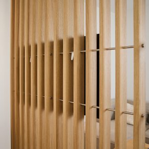 WoodUpp houten lamellenwand met slanke eikenhouten verticale lamellen, bevestigd door horizontale roestvrijstalen stangen en kleine metalen bevestigingsclips. Warme houtnerf, gelijkmatige tussenruimte en subtiele scharnierhaken; zicht op televisiescherm en bank aan de andere zijde, een minimalistische, functionele ruimteafscheiding, natuurkleurige afwerking en fijn afgewerkt oppervlak.