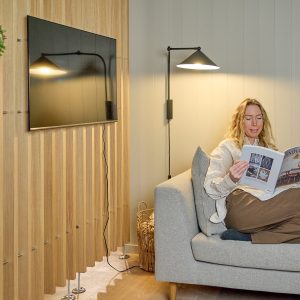 Zithoek met een vrouw op een grijze bank, wandlamp en tv; opvallend zijn de WoodUpp houten lamellenpanelen als sierwand naast de tv, smalle verticale houten latten met zichtbare bevestigingsvoetjes en kabelgeleiding. De warme, natuurlijke WoodUpp-wandbekleding geeft de ruimte textuur en gezellige sfeer.