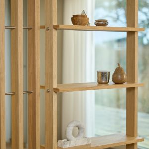 WoodUpp eiken roomdivider/boekenrek met slanke verticale stijlen, verstelbare planken en roestvrijstalen stangen. Afgewerkt met warme olie voor een natuurlijke uitstraling. Op de planken kleine houten schaaltjes, een glanzend metalen bekertje, een peer‑vormig houten doosje en een marmeren ringbeeldje als decoratieve accenten.