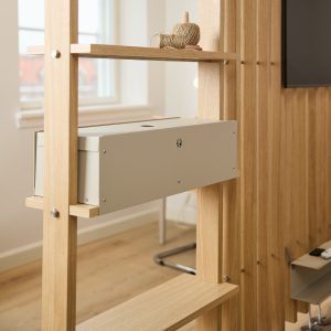 WoodUpp houten roomdivider met lichte eiken latten en geïntegreerde planken; op een plank een klos jute touw en tussen de stijlen een lichtgrijze metalen kabelbox/opbergvak bevestigd. Strakke, minimalistische afwerking met zichtbare RVS-bouten toont functioneel kabelbeheer, ideaal voor kantoor of woonruimte.