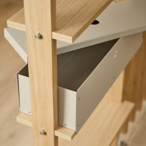 Close-up van een WoodUpp rek: licht eikenhouten stijlen en planken met zichtbare schroeven, gecombineerd met een beige metalen WoodUpp opbergbox met afneembare klep en ronde kabeldoorvoering. Strak, minimalistisch ontwerp met matte afwerking en praktische opbergruimte voor thuis of kantoor, dagelijks gebruik.