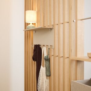WoodUpp houten lattenwand van licht eiken als roomdivider met een beige metalen wandkapstok van WoodUpp: smal bovenplankje met vier geïntegreerde haken. Aan de haken hangen een donkerbruine sjaal, een katoenen netzak en een opgevouwen groene paraplu; een kleine lamp staat op de plank.
