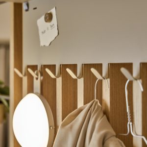 WoodUpp wandpaneel met smalle eiken latten in naturel houtkleur en een rij witte metalen haken; aan de haken hangen een ronde LED-lamp en een jas aan een metalen kleerhanger. Bovenaan houdt een klein WoodUpp houten magneetje een briefje tegen het paneel.