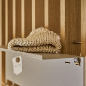 Close-up van een WoodUpp eiken lamellenwand met een WoodUpp matgrijze metalen opbergbox/shelf waarop een opgevouwen crème gebreide trui ligt; een zilverkleurige magneetclip en messing haak zijn zichtbaar tussen de warme houtnerven, en strakke afwerking en subtiele contrasten in licht en materiaal.
