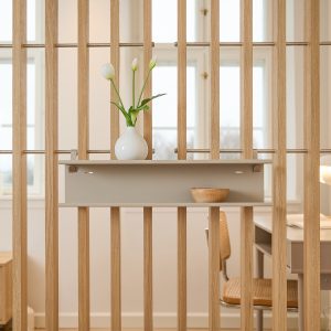 WoodUpp houten lamellenwand van licht eiken met stalen verbindingsstangen, afgewerkt met een beige WoodUpp modulaire wandplank. Op de plank staat een witte keramische vaas met drie witte tulpen en een klein houten schaaltje; op de achtergrond zichtbaar een bureau en rieten stoel.