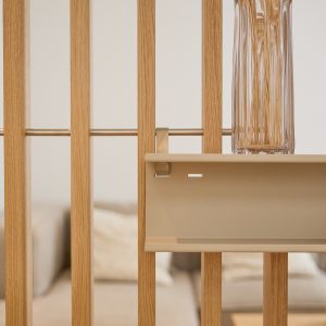 Close-up van een WoodUpp roomdivider met verticale eiken lamellen, waaraan een beige metalen WoodUpp hangplank hangt aan een dunne stang. Op de plank staat een hoog geribbeld glazen vaas met gedroogde stengels; op de achtergrond vervaagde bank en kussens en minimalistische sfeer.