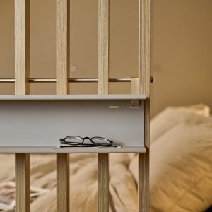WoodUpp-ruimteverdeler van licht eiken met verticale latten, voorzien van een smal grijs minimalistisch design opbergplankje met metalen bevestigingsrail en messing haak. Op het WoodUpp-plankje liggen een bril en een klein notitiepapiertje; op de achtergrond een zacht, warm gekleurde, sfeervolle slaapkamer.