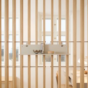 WoodUpp-roomdivider van lichte eikenhouten lamellen met roestvrijstalen staven, gecombineerd met een beige WoodUpp-opbergpaneel en smalle plank die aan de staven hangt. Op de plank staan een metalen kom, een slanke metalen vaas en een klein houten schaaltje in een lichte, minimalistische interieursetting.