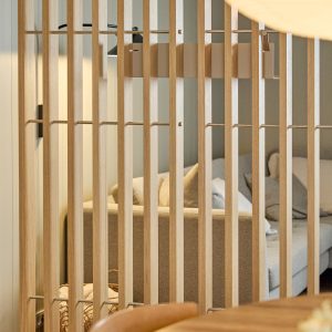 WoodUpp lamellenwand van licht eiken scheidt woonkamer en eetgedeelte; smalle verticale houten lamellen verbonden met dunne metalen stangen. Achter de wand zicht op een grijs stoffen bank en sierkussens. Close-up toont natuurlijke houtnerf, subtiele metalen details en elegante, moderne afwerking van WoodUpp.