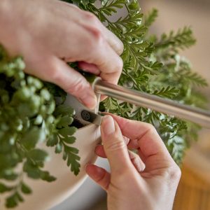 Close-up van handen die een WoodUpp ophangsysteem monteren: een slanke metalen stang in een kunststof montageclip aan een witte hangpot met groen, varenachtig blad. Duidelijk zichtbaar zijn de WoodUpp metalen stang en de bijbehorende montageclip die de bloempot veilig vastzet.