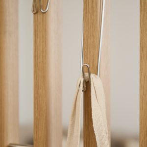Close-up van WoodUpp houten lattenpaneel met lichte eikennerf en natuurlijke afwerking, gecombineerd met slanke verchroomde metalen haken die om de latten klemmen; één haak draagt een beige geweven draagriem. Minimalistische, functionele WoodUpp opberghaken en latten voor ruimtebesparend ophangen van accessoires.