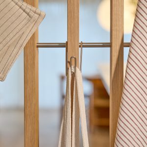 WoodUpp houten wandpaneel met smalle eikenlatten en een horizontale roestvrijstalen roede, waaraan twee metalen S-/U-haken van WoodUpp hangen. Aan de haken hangen een crèmekleurige katoenen draagband en gestreepte theedoeken, met een natuurlijke warme houttint en strak geborsteld metaal, geschikt voor keukens en entrees.
