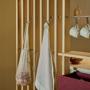 WoodUpp eiken lattenwand met horizontale RVS-rails en bijpassende metalen haakjes waarop een gestreepte theedoek en een katoenen netzak hangen. Ook zichtbaar: een ingebouwde WoodUpp plank, metalen mokhangers met witte mokken en een rood vilten opbergbox, alles in warme, natuurlijke afwerking.