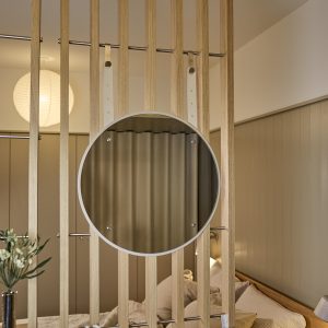 WoodUpp houten lattenwand functioneert als roomdivider; vooraan hangt een WoodUpp ronde spiegel met smalle witte rand en glanzende spiegelruit, bevestigd aan verstelbare crèmekleurige leren banden die aan metalen stangen en zichtbare bevestigingsschroeven hangen. De lichte eiken latten creëren een warme, minimalistische slaapkamerafscheiding.