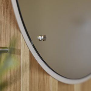 WoodUpp ronde spiegel met matte witte rand, subtiele zwarte binnelijst en zichtbare RVS-afstandhouder, bevestigd op een WoodUpp eikenhouten lattenpaneel met strakke afwerking. Close-up toont het heldere spiegeloppervlak, het metalen bevestigingspunt en een onscherpe groene plant op de voorgrond in warme, natuurlijke tinten.