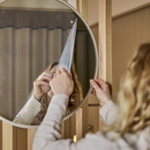 Close-up van iemand die de beschermfolie van een WoodUpp ronde spiegel verwijdert, bevestigd in een lichtgrijze metalen rand en gemonteerd op een WoodUpp houten lamellenwand. De reflectie toont handen en golvend haar, warme houtkleur, strakke, minimalistische uitstraling en zachte verlichting.