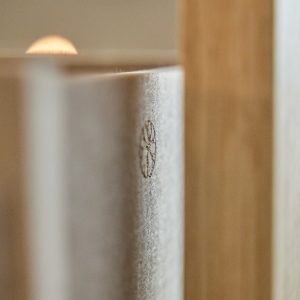 Close-up van een WoodUpp-beeld: de rand van een grijs vilten akoestisch paneel met een geperst WoodUpp-logo zichtbaar in het oppervlak, naast een lichte houten lat of frame. Toont producten van WoodUpp: vilten paneel en houten lat; zachte achtergrondverlichting benadrukt textuur.