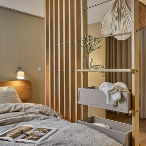 WoodUpp houten scheidingswand met smalle eiken lamellen, metalen kledingstang en ingebouwde planken met zachte, grijze opbergboxen (twee niveaus), zichtbaar met een opgevouwen trui. Op een plank staan een kleine vaas met groen, parfumflacon en glazen schaaltje; sfeervol naast bed en lamp.