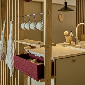 WoodUpp eiken roomdivider met open houten frame en rek met metalen haken waarop vijf witte mokken hangen; een ingebouwde bordeauxrode vilten lade/organizer schuift onder het werkblad. Minimalistische keuken met houten blad, kraan en ophanghaken voor theedoeken, warm verlicht en compacte opbergruimte voor keukenaccessoires.