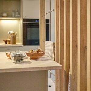 WoodUpp houten slatwand en bijbehorend massief houten werkblad in een moderne keuken: verticale eiken latten met geïntegreerde roestvrijstalen handdoekstangen, lichtkleurig keukenblad met houten fruitkom en glazen pot, subtiele verlichting, natuurlijke nerven en warme tinten, minimalistische uitstraling.