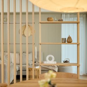 WoodUpp scheidingswand van slank eiken met verticale latten en open planken; op de planken een houten schaaltje, glazen piramide, metalen potje en houten peerdecoratie, plus een marmeren ringbeeld en brandende dinerkaars, met zicht op lichte zithoek en houten eettafel op de achtergrond.