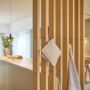 WoodUpp houten lamellenwand van warme eikenhouten verticale latten met slanke roestvaststalen roedes als haken. Aan de ingebouwde roedes hangen een crème-gestreepte pannenlap en bijpassende theedoek. Strak, natuurlijke afwerking, subtiele roomdivider naast keukenblad met zacht licht en woondecoratie op de achtergrond.