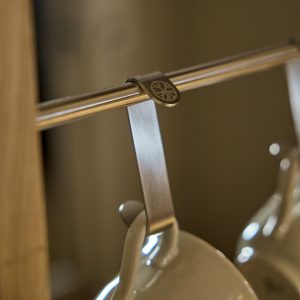 Close-up van een WoodUpp-montage: een metalen hangrail met een geborstelde roestvrijstalen WoodUpp-kopjeshaak waaraan een crèmekleurig keramisch mokje hangt, en een klein metalen WoodUpp-clipje met rond, uitgesneden boomlogo bevestigd aan de buis, met satijnachtige afwerking.
