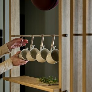 WoodUpp houten roomdivider met licht eiken open plank en geïntegreerde metalen ophangrail met vijf zilverkleurige mokhaken; vier crème keramische mokken hangen eraan terwijl iemands handen één mok vastpakken. Op de plank ligt een klein takje kruiden. Functionele, minimalistische keukenopstelling van WoodUpp.