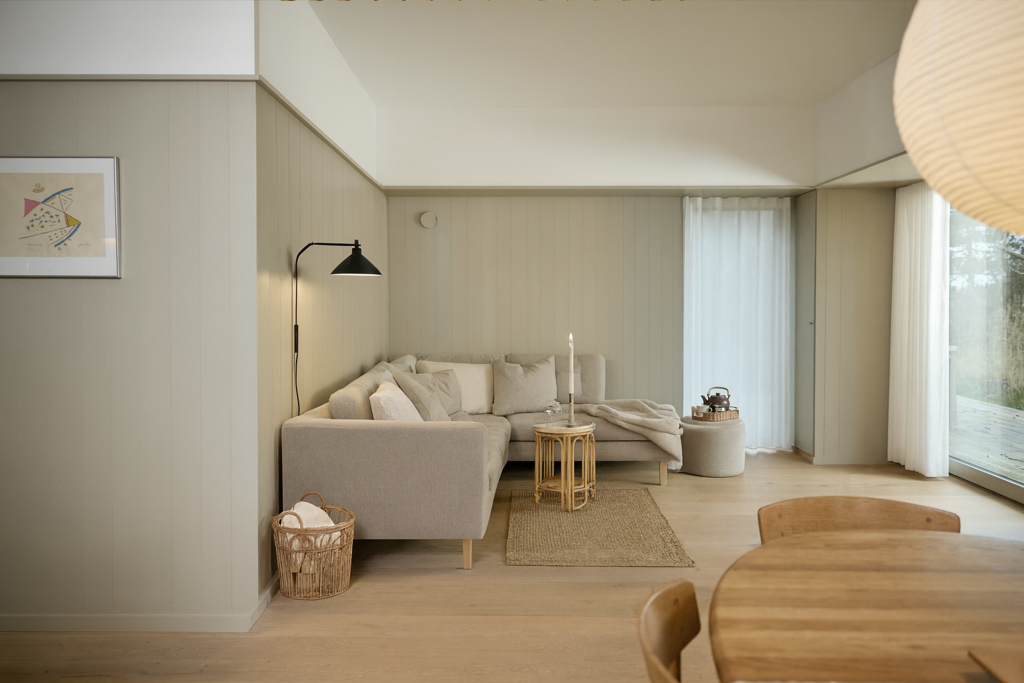 Een sfeervolle Scandinavische woonkamer en eethoek met een beige hoekbank en houten tafel. De wanden zijn bekleed met WoodUpp wandpanelen. Deze panelen zijn verticaal geplaatste, beige geschilderde brede planken die een rustige, gestructureerde achtergrond vormen voor het lichte interieur.