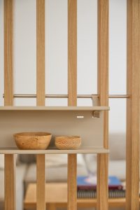 WoodUpp roomdivider van slanke, licht eikenhouten latten met een beige, matte metalen plankje van WoodUpp, bevestigd door een subtiele beugel. Op het plankje staan twee handgedraaide houten kommetjes — één groter, één kleiner — met zichtbare nerven en warme houttekening, en vormt een subtiele roomdivider in een modern interieur.