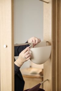 Handen bevestigen een beige WoodUpp hangende opbergpot aan een verchroomde stang binnen een licht eiken houten frame; op de onderste stang is een bordeaux WoodUpp tas-/accessoirehouder zichtbaar. Minimalistische, matte kunststof pot met eenvoudige klem voor gemakkelijk ophangen en organiseren, functioneel, ruimtebesparend en sfeervol.