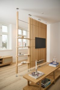 WoodUpp roomdivider van licht eiken met verticale lamellen, plafond- en vloerbevestigde roestvrijstalen stangen, geïntegreerde tv-bevestiging en open planken; daarnaast bijpassende WoodUpp eiken salontafel en dressoir met gevlochten deurfront, natuurlijke afwerking en verstelbare planken voor decoratieve objecten, Scandinavische uitstraling.