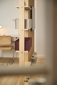 WoodUpp houten roomdivider met strakke, natuurlijke afwerking, verticale eikenlatten en zichtbare schroefverbindingen, uitgerust met twee WoodUpp opbergboxen: een grijze en een bordeauxrode vilten box, plus kleine houten plankjes; op de achtergrond een werkhoek met bureau en rieten stoel vaag zichtbaar.