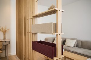 WoodUpp houten ruimteverdeler met een slank eiken frame en twee opgehangen vilten opbergbakken — één beige en één bordeaux — geplaatst op houten schappen. Strak, licht en modern interieur met subtiele decoratie, bank en bureau zichtbaar op de achtergrond in de woonkamer.